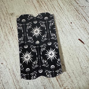 May include: Black and white bookmark with a celestial design. The bookmark features a repeating pattern of suns, crescent moons, and leafy branches. The edges are scalloped.