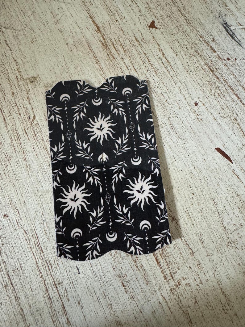 May include: Black and white bookmark with a celestial design. The bookmark features a repeating pattern of suns, crescent moons, and leafy branches. The edges are scalloped.
