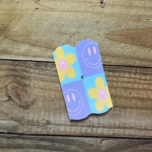 May include: A small, folded paper card with a repeating pattern of yellow flowers and smiling faces on a light blue and purple background. The card is set against a wooden surface.