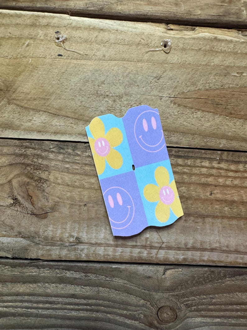 May include: A small, folded paper card with a repeating pattern of yellow flowers and smiling faces on a light blue and purple background. The card is set against a wooden surface.