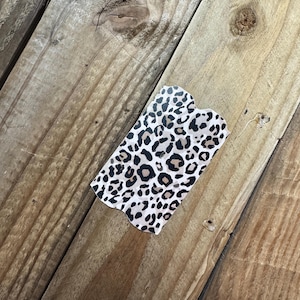 K&ouml;nnte beinhalten: Ein rechteckiger Artikel mit Leopardenmuster und gewelltem Rand. Der Druck zeigt schwarze und braune Flecken auf cremefarbenem Hintergrund. Er liegt auf einer verwitterten Holzoberfl&auml;che.