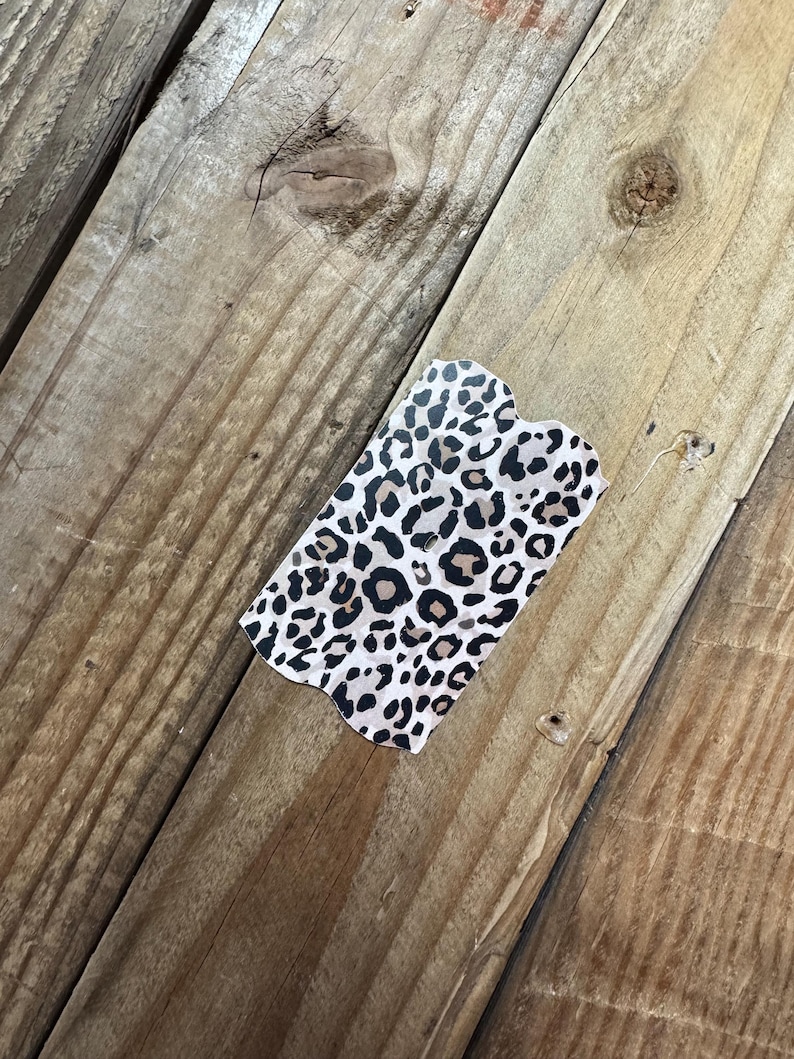 K&ouml;nnte beinhalten: Ein rechteckiger Artikel mit Leopardenmuster und gewelltem Rand. Der Druck zeigt schwarze und braune Flecken auf cremefarbenem Hintergrund. Er liegt auf einer verwitterten Holzoberfl&auml;che.