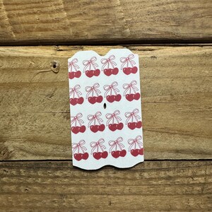 May include: A white tag with a scalloped edge featuring a repeating pattern of red cherries with green bows. The tag is made of cardstock and is set against a wooden background.