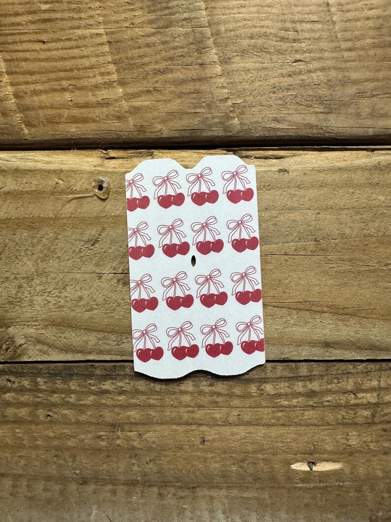 May include: A white tag with a scalloped edge featuring a repeating pattern of red cherries with green bows. The tag is made of cardstock and is set against a wooden background.