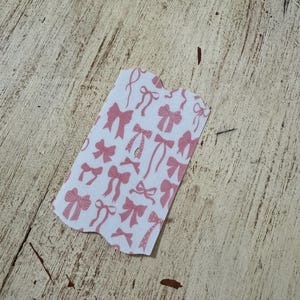 Op de afbeelding: Een klein, onregelmatig gevormd stuk papier met een witte achtergrond en een patroon van roze strikken. De strikken vari&euml;ren in stijl en grootte, sommige hebben linten en andere lijken op eenvoudige knopen. Het papier ligt op een lichtgekleurd, getextureerd houten oppervlak.