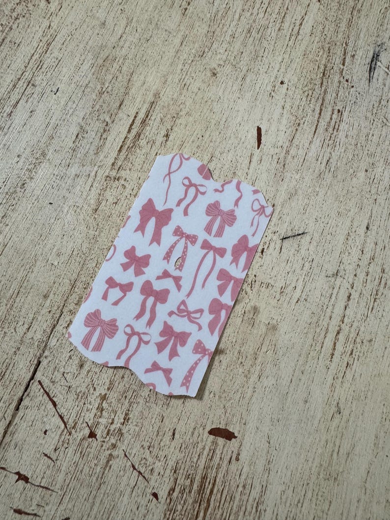 Op de afbeelding: Een klein, onregelmatig gevormd stuk papier met een witte achtergrond en een patroon van roze strikken. De strikken vari&euml;ren in stijl en grootte, sommige hebben linten en andere lijken op eenvoudige knopen. Het papier ligt op een lichtgekleurd, getextureerd houten oppervlak.