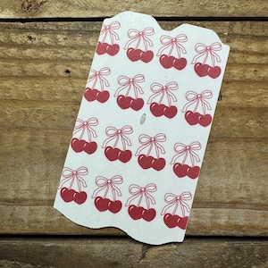 May include: A white tag with a repeating pattern of red cherries and bows. The cherries are in pairs, each with a stem and a bow at the top. The tag has a scalloped edge and is set against a wooden background.