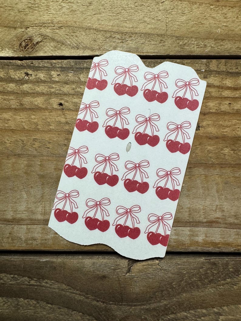 May include: A white tag with a repeating pattern of red cherries and bows. The cherries are in pairs, each with a stem and a bow at the top. The tag has a scalloped edge and is set against a wooden background.