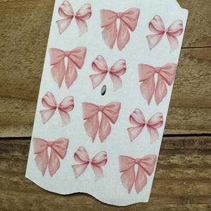 May include: A white tag with a scalloped top edge features multiple illustrations of pink bows. The bows are in various sizes and orientations, with detailed shading. The tag has a small hole near the center.