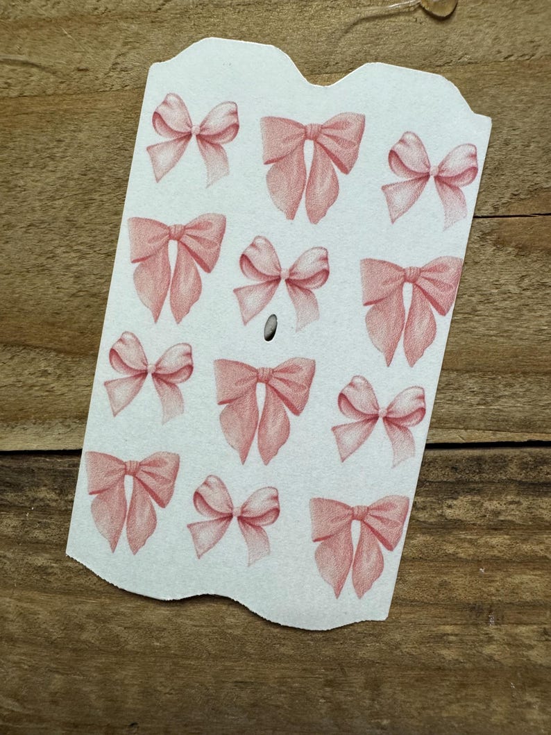 May include: A white tag with a scalloped top edge features multiple illustrations of pink bows. The bows are in various sizes and orientations, with detailed shading. The tag has a small hole near the center.