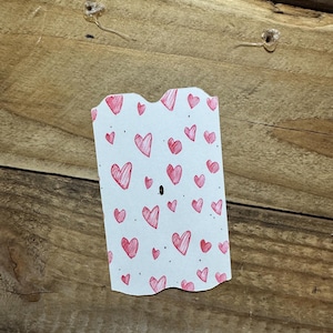 Puede incluir: Una tarjeta blanca con un borde superior festoneado, cubierta de peque&ntilde;os corazones rojos dibujados a mano. La tarjeta est&aacute; colocada sobre una superficie de madera, con dos chinchetas transparentes visibles. Los corazones var&iacute;an en tama&ntilde;o y est&aacute;n dispersos por la tarjeta.