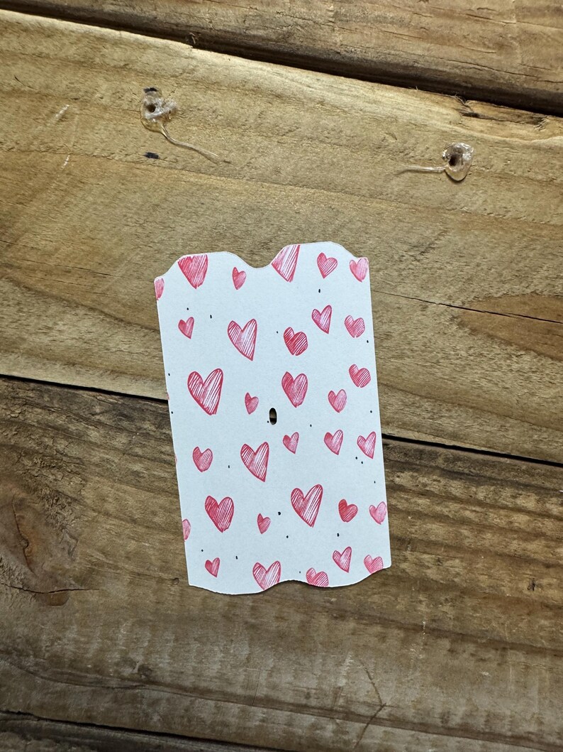 Puede incluir: Una tarjeta blanca con un borde superior festoneado, cubierta de peque&ntilde;os corazones rojos dibujados a mano. La tarjeta est&aacute; colocada sobre una superficie de madera, con dos chinchetas transparentes visibles. Los corazones var&iacute;an en tama&ntilde;o y est&aacute;n dispersos por la tarjeta.