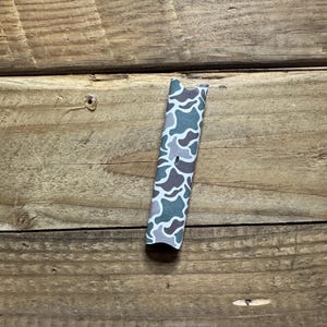 Puede incluir: Un objeto rectangular con un patr&oacute;n de camuflaje en tonos verdes, grises y marrones. Se encuentra verticalmente sobre un fondo de madera. El dise&ntilde;o de camuflaje presenta formas y patrones irregulares. Mide unos 10 cm de largo.