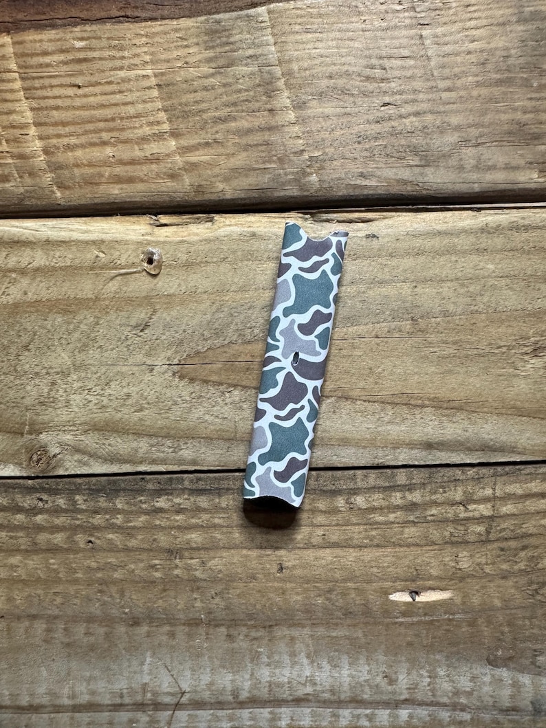 Puede incluir: Un objeto rectangular con un patr&oacute;n de camuflaje en tonos verdes, grises y marrones. Se encuentra verticalmente sobre un fondo de madera. El dise&ntilde;o de camuflaje presenta formas y patrones irregulares. Mide unos 10 cm de largo.