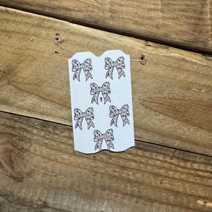 May include: A light blue paper tag with six black bow illustrations. The bows have a dotted pattern. The tag is on a wooden surface with a diagonal grain.