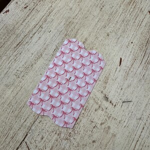 Puede incluir: Una peque&ntilde;a etiqueta con un dise&ntilde;o de lazo rosa y blanco. La etiqueta tiene un borde festoneado y est&aacute; colocada sobre una superficie de madera clara y desgastada.