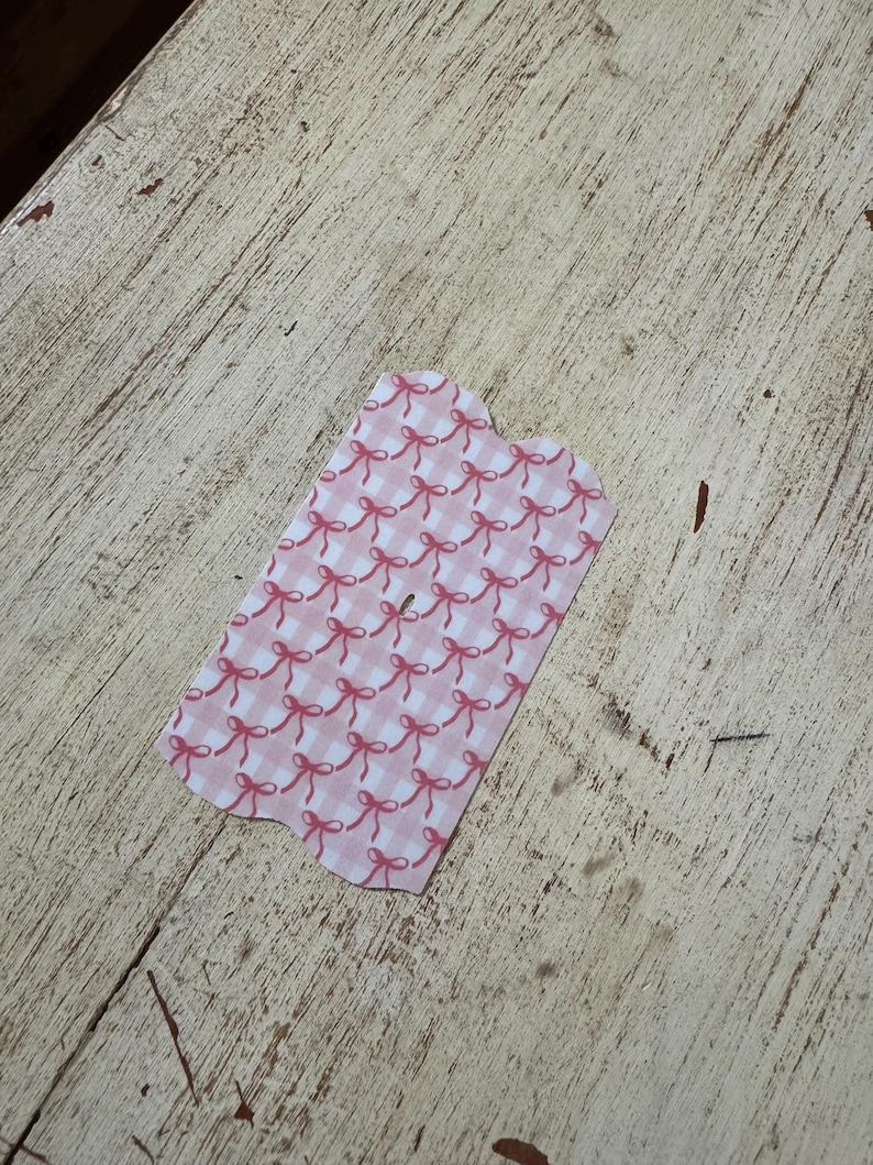 Puede incluir: Una peque&ntilde;a etiqueta con un dise&ntilde;o de lazo rosa y blanco. La etiqueta tiene un borde festoneado y est&aacute; colocada sobre una superficie de madera clara y desgastada.