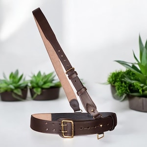 May include: A dark brown leather belt with a matching shoulder strap. The belt has a large brass buckle and adjustment holes. The strap is attached with brass hardware. Potted plants are in the background.