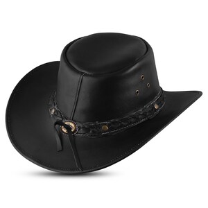 Op de afbeelding: Een zwarte leren cowboyhoed met een gevlochten band en een decoratieve gesp. De hoed heeft een brede rand en een hoge kroon.