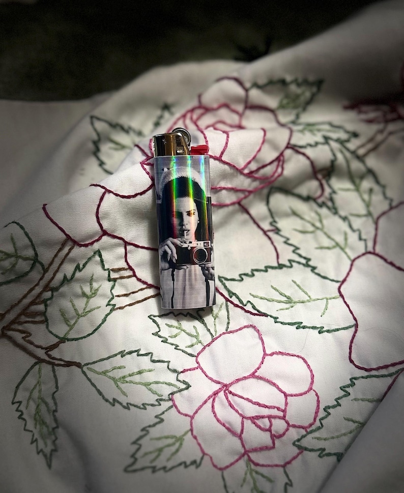 May include: A clear, rectangular lighter with a gold-colored top and a red ignition button. The lighter features a black and white image of a person holding a camera. The lighter is resting on a white fabric with embroidered pink roses and green leaves.