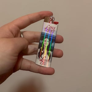 May include: A silver lighter featuring a portrait of a woman with long dark hair and the text "Lana Del Rey" and "Lust for Life". The lighter has a red ignition button and a silver metal top.