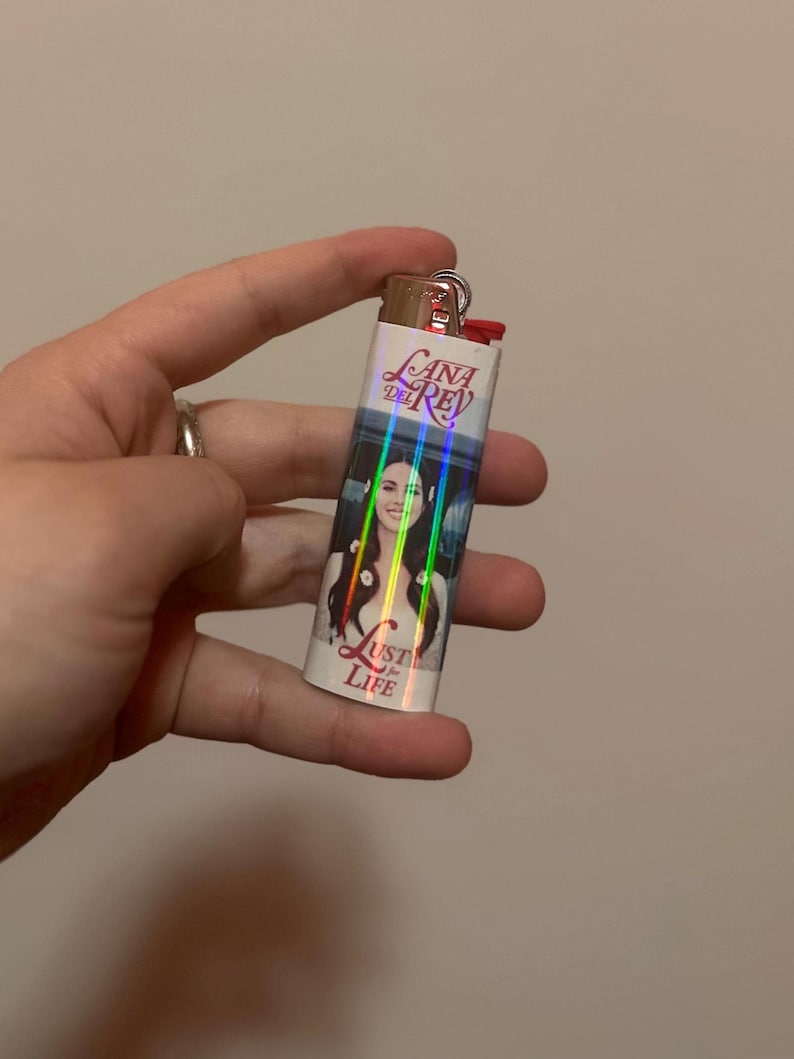 May include: A silver lighter featuring a portrait of a woman with long dark hair and the text "Lana Del Rey" and "Lust for Life". The lighter has a red ignition button and a silver metal top.