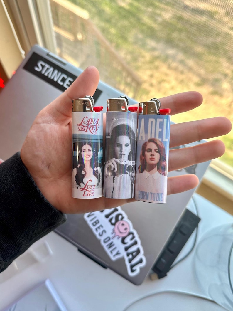 May include: Three white lighters with images of Lana Del Rey. Each lighter features a different album cover design, with text including "Lust for Life" and "Born to Die". The lighters have red ignition buttons.