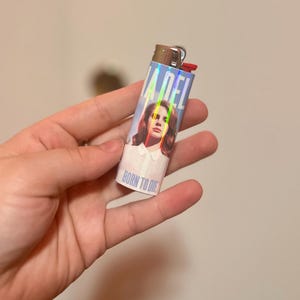 May include: A rectangular lighter with a holographic design featuring the text "LANA DEL" and the words "BORN TO DIE". The lighter has a red ignition button and a silver metal top. The image on the lighter is of a woman with brown hair.