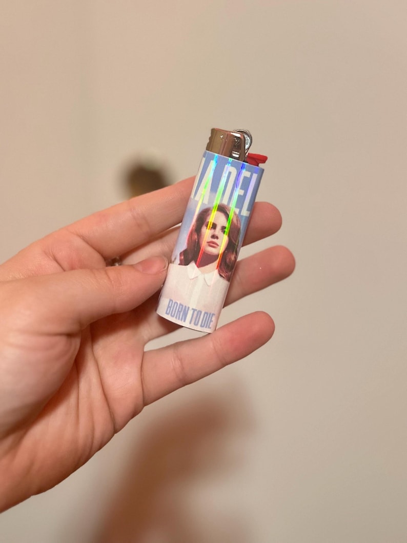 May include: A rectangular lighter with a holographic design featuring the text "LANA DEL" and the words "BORN TO DIE". The lighter has a red ignition button and a silver metal top. The image on the lighter is of a woman with brown hair.