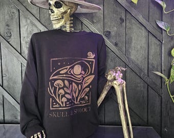Puede incluir: Una sudadera negra con un gráfico color cobre de una calavera, plantas y las palabras "SKULL & SPROUT". La sudadera la lleva un esqueleto con un sombrero gris. El fondo es una pared de madera oscura con las palabras "& Sprout" en dorado.
