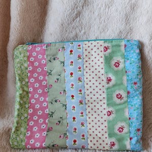 May include: A patchwork zipped pouch featuring vertical fabric strips in floral patterns and colours, including pink, green, and blue. The pouch has a teal zip and is likely handmade.