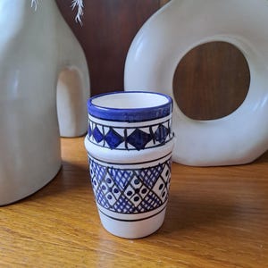 May include: A white ceramic cup with a blue and black geometric pattern. The cup is decorated with a repeating diamond pattern and small circles.