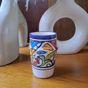 May include: A white ceramic cup with a blue rim and colorful floral pattern. The cup is decorated with yellow, orange, red, blue, and green floral designs.