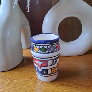 May include: A white ceramic cup with a blue rim and colorful geometric designs. The cup is decorated with red, yellow, green, and blue geometric shapes.