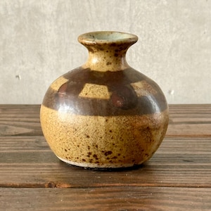 May include: A small, round ceramic vase with a narrow neck and a speckled, textured surface. The vase features a brown geometric pattern around the middle and a light brown base. The vase is displayed on a wooden surface.
