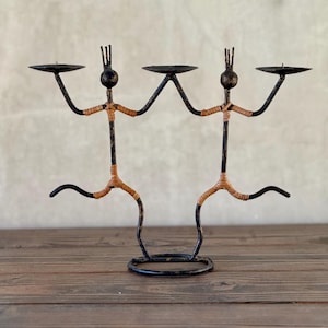 May include: A black metal candle holder with a unique stick figure design. The figures have outstretched arms holding candle platforms. The legs are bent, and the bodies are wrapped with tan-colored material. The candle holder sits on a round base.