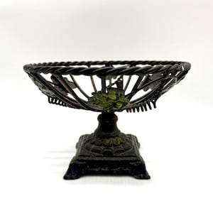 May include: A black metal bowl with a twisted wire design and green leaf accents sits on a square base.