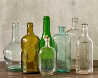 May include: A collection of antique glass bottles in various shapes and colors, including clear, green, and amber. Some bottles have labels or embossed details. The bottles are arranged on a wooden surface.