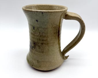 May include: A handmade stoneware mug with a unique, organic shape. The mug features a wide, flared rim and a sturdy handle. The exterior has a mottled, earthy color palette of greens and browns, with subtle variations in texture.