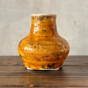 May include: A small, handcrafted ceramic vase with a rounded body and a narrow neck. The vase has a warm, golden-brown color with darker brown speckles and horizontal texture. The base is white.