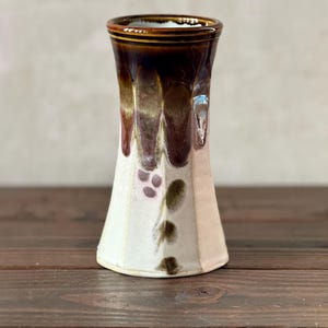 May include: A handmade ceramic vase with a unique design. The vase features a white base with brown and purple accents. The top rim is a dark brown color. The vase has a flared shape and is suitable for displaying flowers or as a decorative piece.
