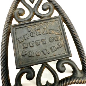 May include: Antique cast iron trivet with a decorative design. The trivet is black with a square center that reads "NEW ENGLAND BUTT CO. PROV. R.I." The trivet has a rope-like border and ornate scroll and heart details.