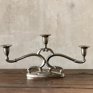 May include: A silver-toned candelabra with three candle holders. The candelabra has a decorative, curved design with a round base. The candle holders extend outwards from a central stem. The background is a neutral gray wall, and the candelabra sits on a wooden surface.
