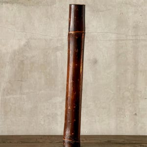 May include: A tall, cylindrical bamboo container with a dark brown finish. The bamboo has a natural, textured surface with visible knots and variations in color. The container is standing upright on a wooden surface.
