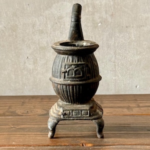 May include: A small, antique cast iron stove with a dark, weathered finish. The stove has a rounded body, a chimney-like top, and sits on three small legs. The design includes decorative details and a door-like feature on the front.