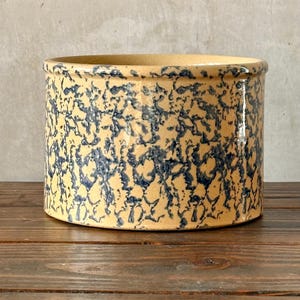 May include: A cylindrical stoneware crock with a beige base and a dark blue, speckled pattern. The rim is slightly rounded. The crock sits on a dark wooden surface. This vintage-style container is suitable for kitchen or home decor.