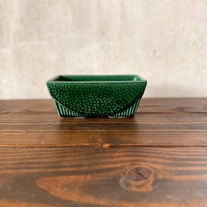 May include: A square, dark green ceramic bowl with a textured, dotted pattern on the upper half and vertical lines on the lower half. The bowl sits on a dark brown wooden surface.
