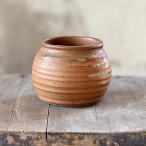 May include: A small, round, terracotta-colored ceramic pot with horizontal ridged detailing. The pot has a wide opening and sits on a weathered wooden surface. The pot's color is a warm, earthy tone, suggesting a rustic or vintage aesthetic.