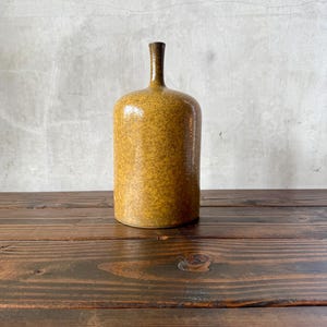 May include: A cylindrical ceramic bottle with a narrow neck and a rounded top. The surface features a mottled, textured mustard yellow and brown glaze with a glossy finish. The bottle has a flat base and stands upright as a single decorative vessel.