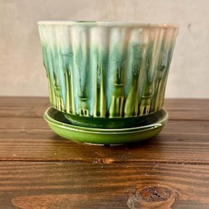 May include: A ceramic planter with a matching saucer. The planter is cylindrical with vertical ridges and a green glaze that drips down the sides. The top rim is white and the saucer is a solid green.